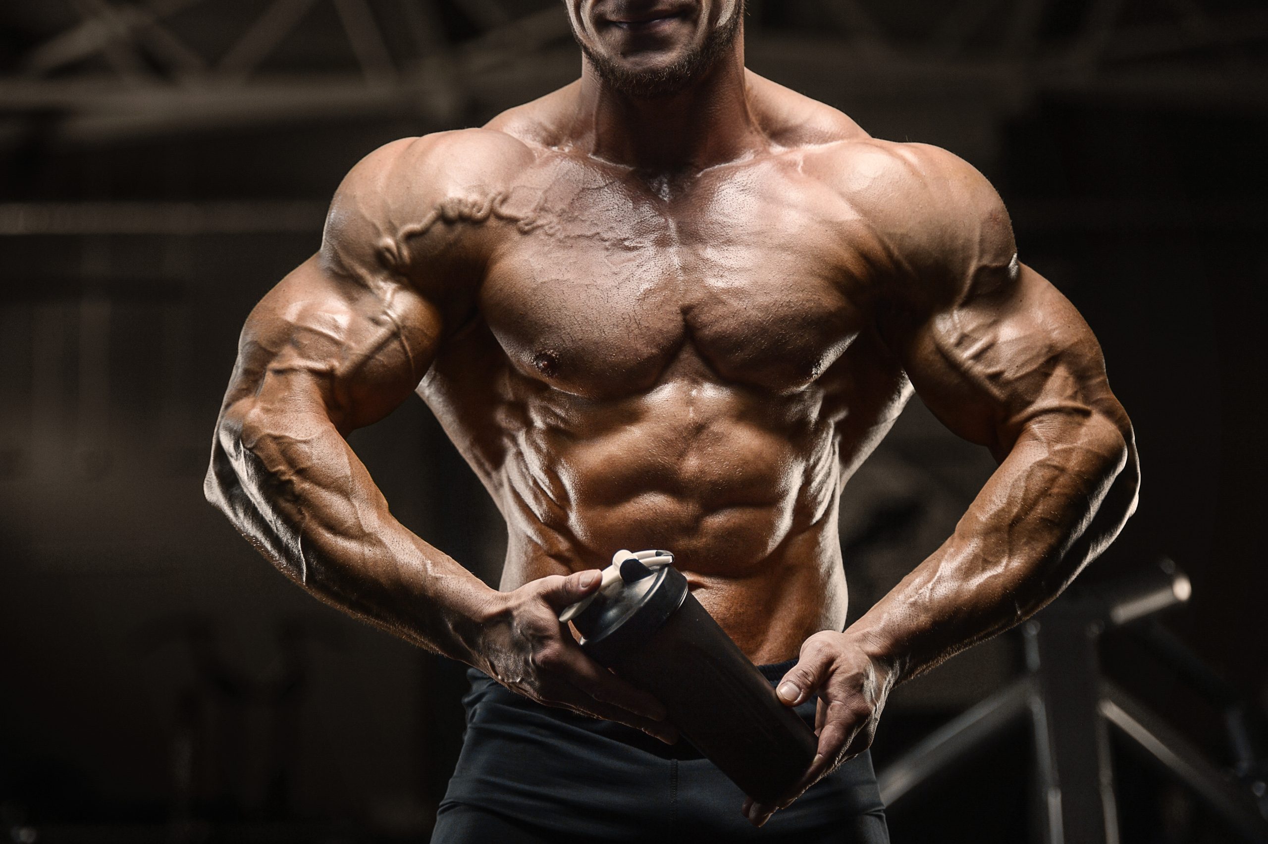A muscular bodybuilder posing in a gym, holding a shaker bottle. Prominent vascularity and bulging veins are visible across his chest, shoulders, and arms.
