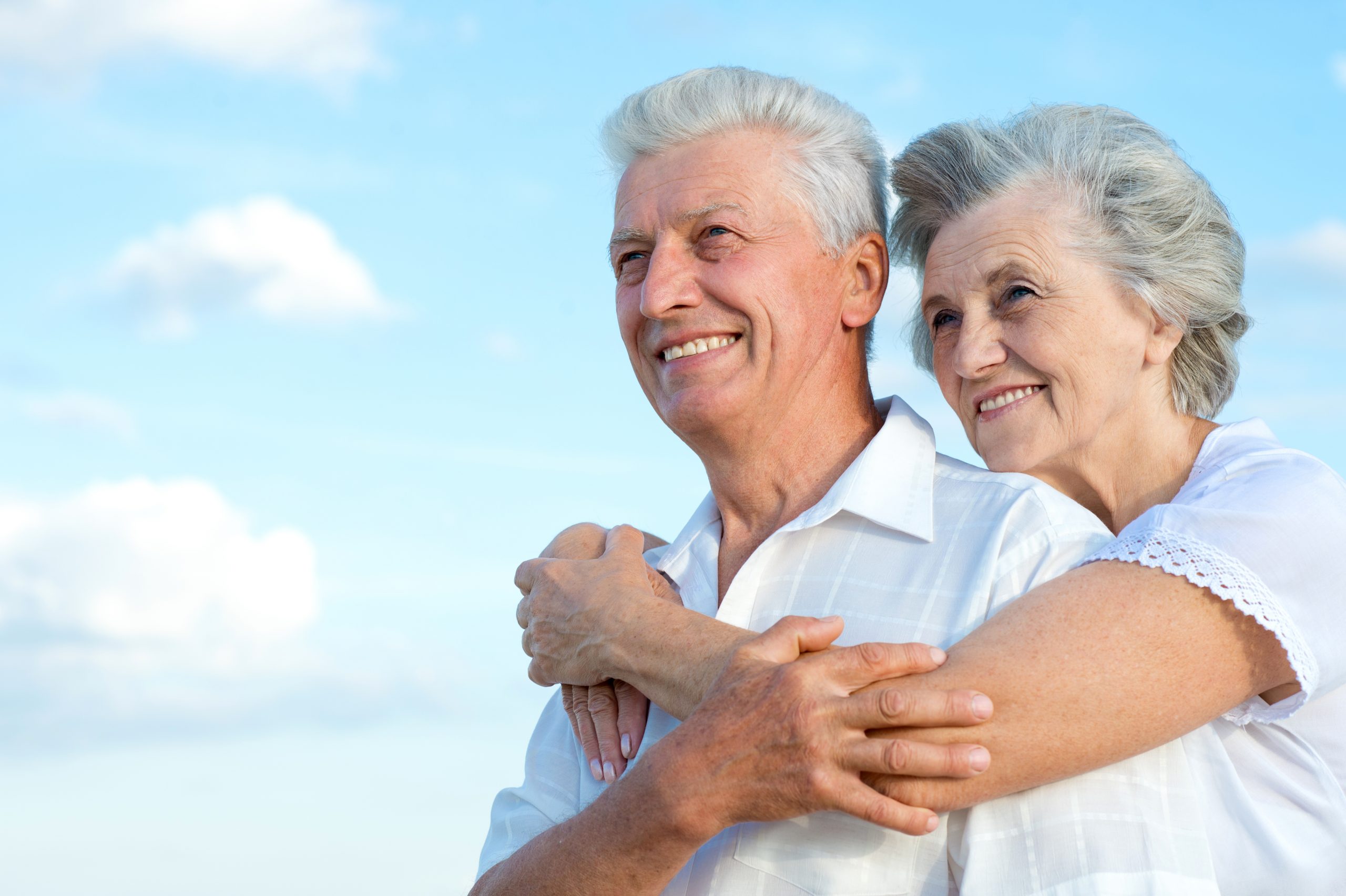 A happy senior couple with grey hair smiling and looking into the distance outdoors. The woman is hugging the man from behind against a clear blue sky with soft white clouds.
