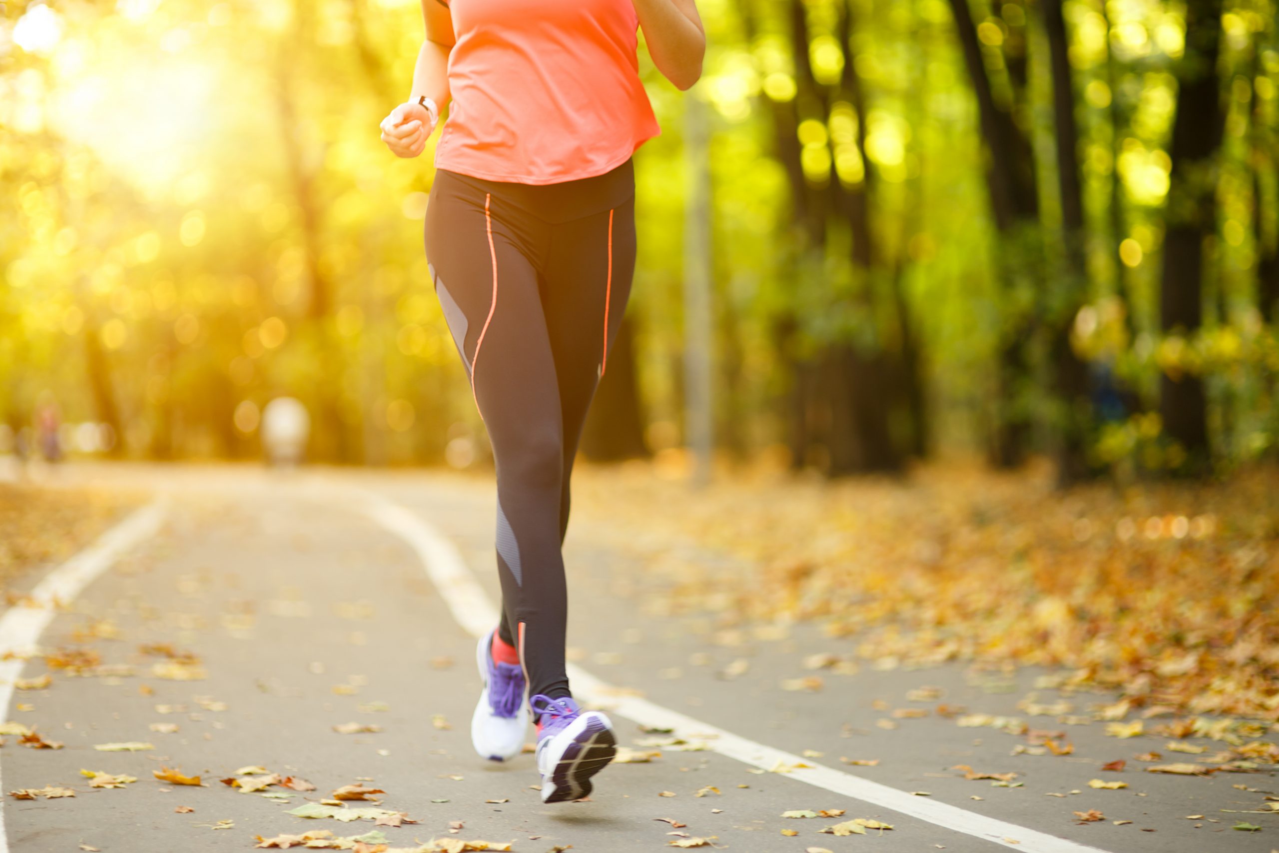 A person wearing an orange shirt and black leggings jogging on a paved park path covered in autumn leaves during a golden sunset.