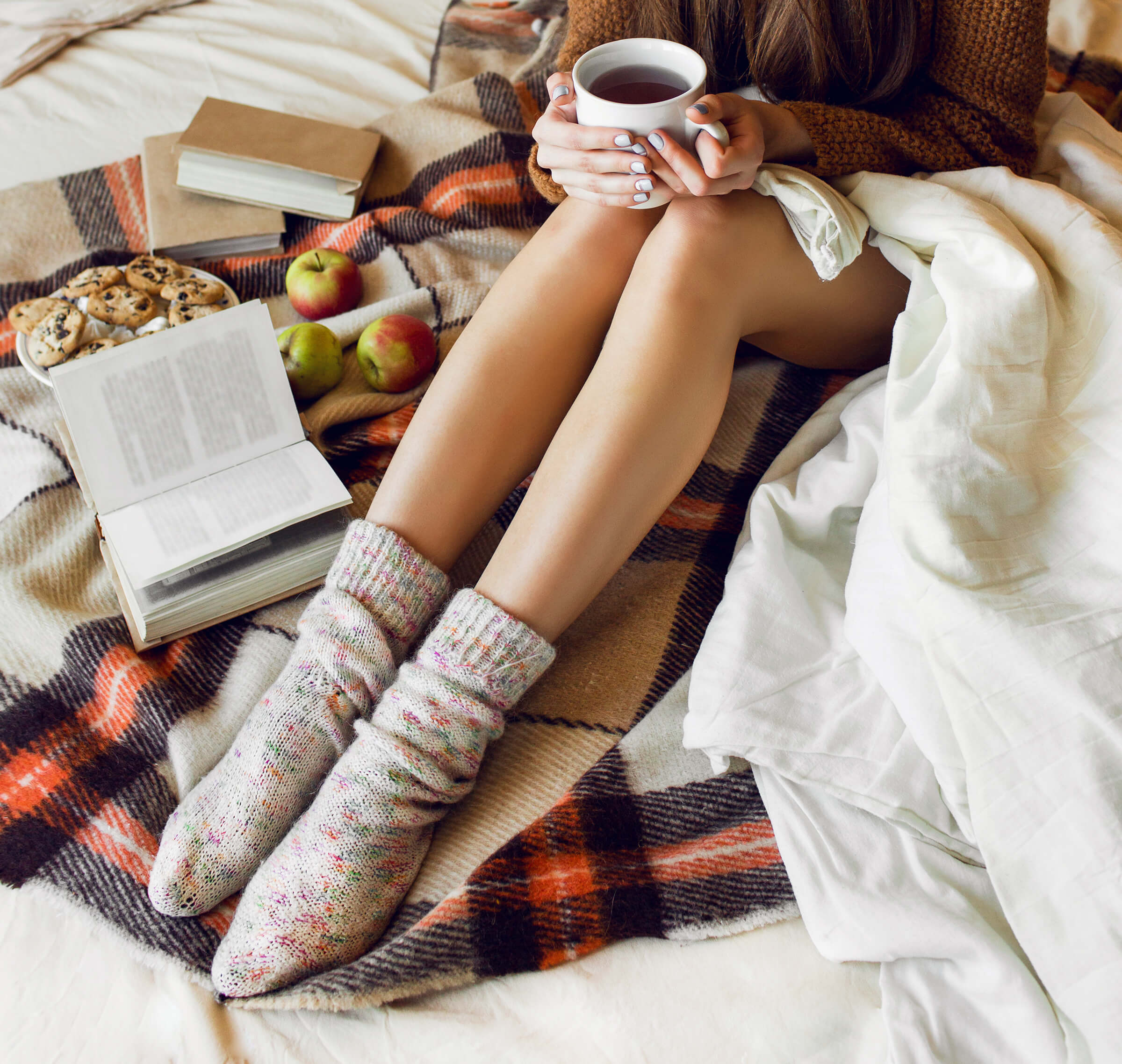 A top-down view of a person wearing cozy speckled wool socks sitting on a plaid blanket, holding a white mug with books, apples, and cookies nearby.