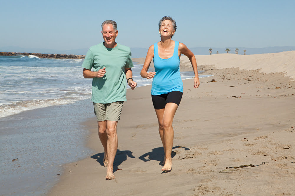 An active senior couple laughing while jogging barefoot along a sandy beach near the ocean.