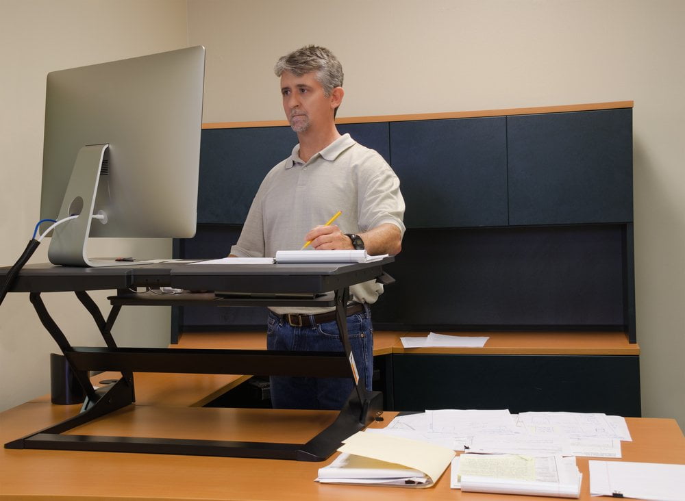 A man in a polo shirt and jeans using a black adjustable standing desk converter while taking notes in an office.