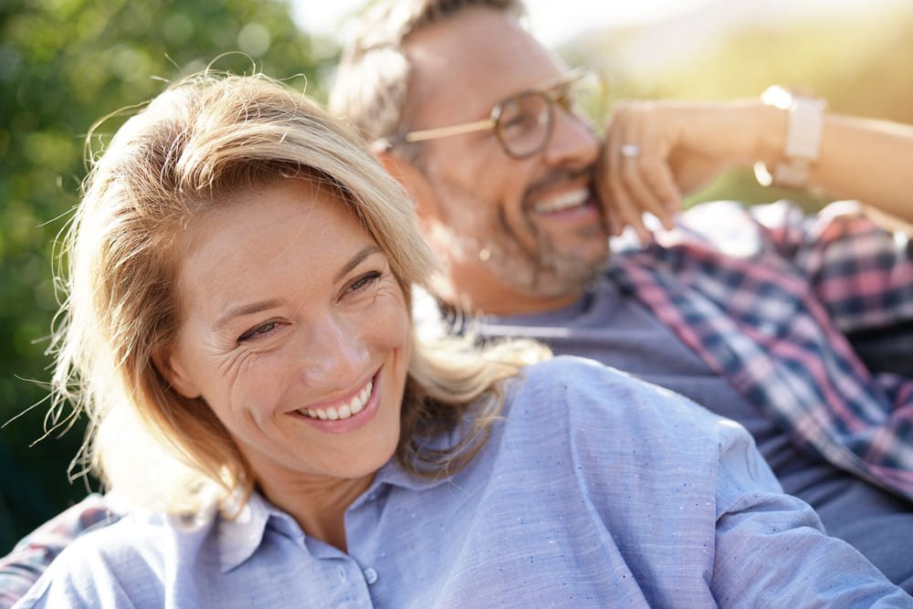 A close-up, candid photograph of a smiling middle-aged couple relaxing outdoors in bright sunlight.