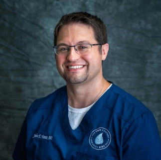 A professional headshot of Dr. John Vance, a medical physician at The Vein Company, smiling in a clinical office setting.