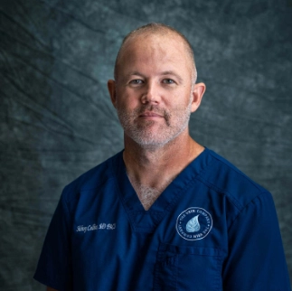 A professional headshot of Dr. Sid Collins, a medical physician at The Vein Company, smiling in a clinical office setting.
