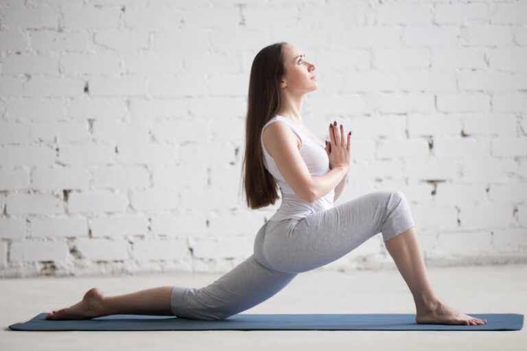 A woman with long dark hair practicing a low lunge yoga pose on a blue mat in front of a white brick wall, demonstrating a lifestyle activity that promotes leg circulation and flexibility.